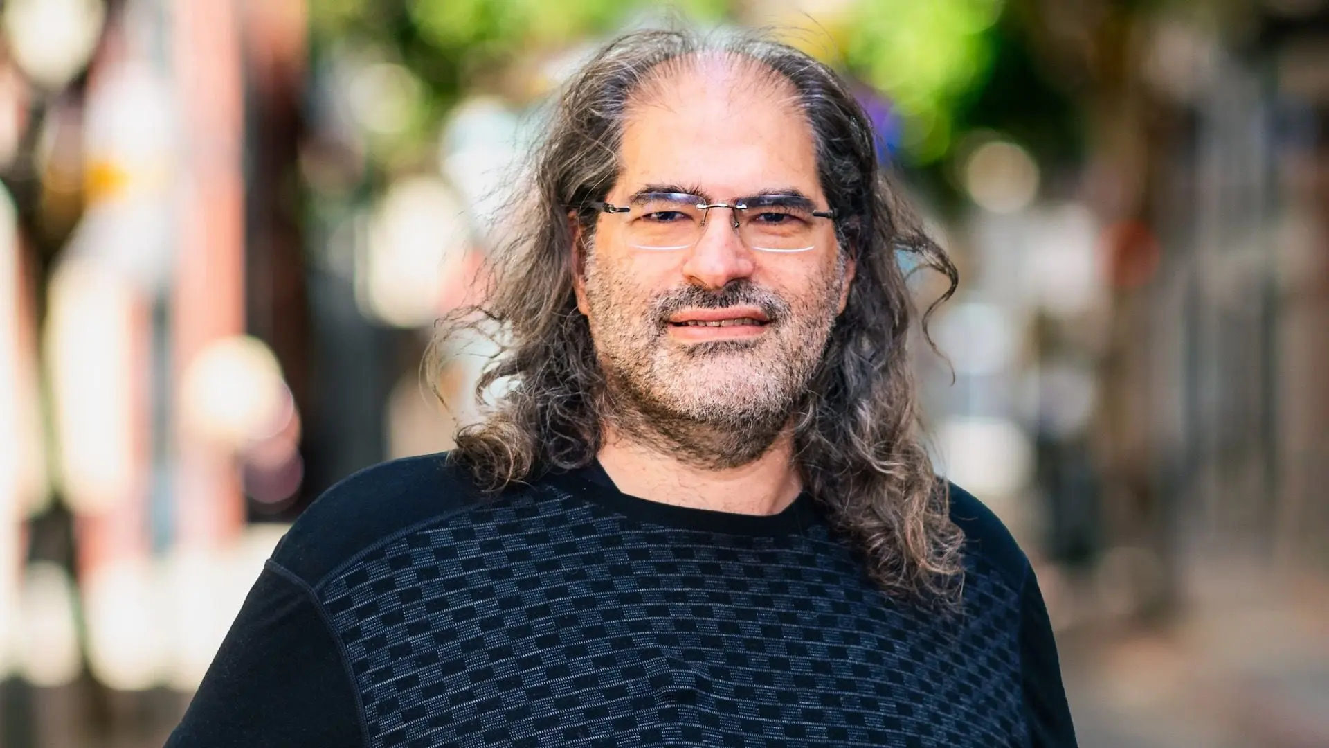 Portrait of a man with long hair and glasses smiling in an outdoor setting.