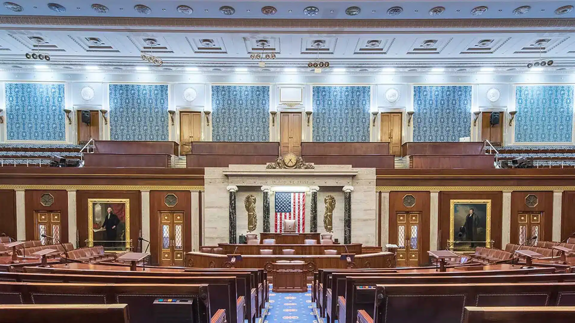 Interior view of the US House chamber with American flag and historical portraits.