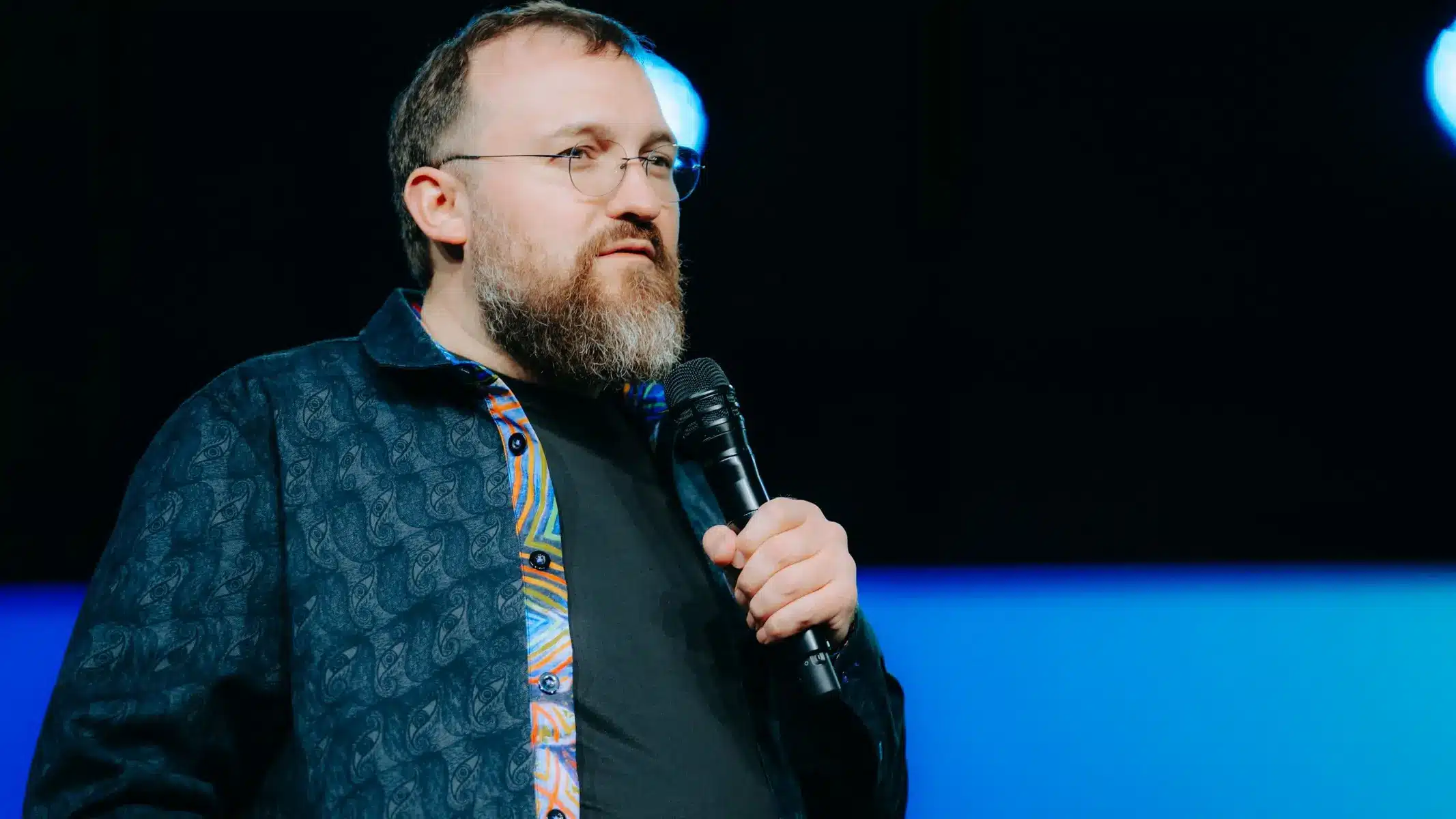 Charles Hoskinson with a beard and glasses holding a microphone during a presentation